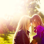Young daughter with glasses and her contemporary mother are touching heads together while holding hands outdoors on a sunny autumn afternoon. Genuine lens flare, real family members and emotions. AdobeRGB profile.