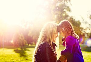 Young daughter with glasses and her contemporary mother are touching heads together while holding hands outdoors on a sunny autumn afternoon. Genuine lens flare, real family members and emotions. AdobeRGB profile.