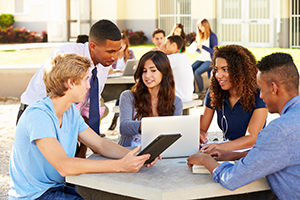 High School Students Working On Campus With Teacher Using Electronics