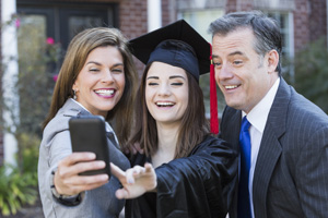 A family celebrating the graduation of their daughter from high school, taking a selfie with a mobile phone.  They are posing in front of their home.  The teenage girl is wearing a black cap and gown with red tassel.  She is standing in the middle between her father, wearing a suit, and mother in a gray jacket.