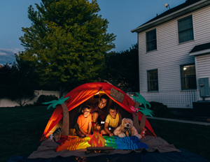 A dad with his two sons camp out in the backyard of their home due to the coronavirus restrictions and quarantine. They have pitched a tent and created a beach scene with stuffed animal friends and have a fake campfire. They are making the best of their situation and long to return to the real outdoors.